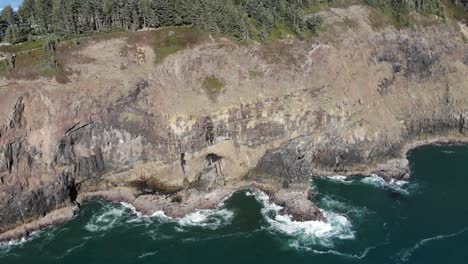 Drone-flies-near-a-cliff-on-the-ocean,-waves-can-be-seen-crashing-onto-the-rocks-below