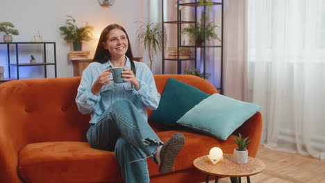atractiva mujer joven sonriente chica bebiendo una taza de café o té de hierbas sentada en el sofá de casa