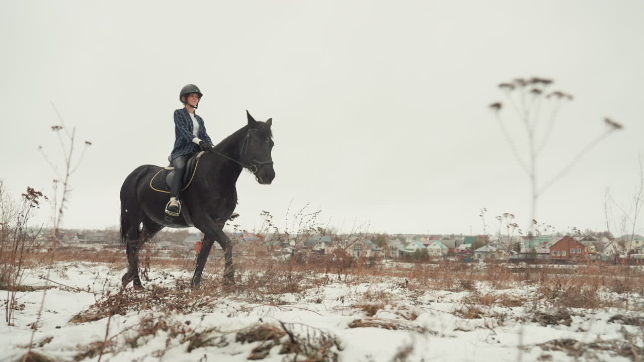 Studente che pratica il controllo durante un lento trotto a cavallo. Coppia che lavora insieme sulle abilità equestri con tempo grigio. Individuo che cavalca un cavallo nero attraverso campi invernali come parte dell'apprendimento