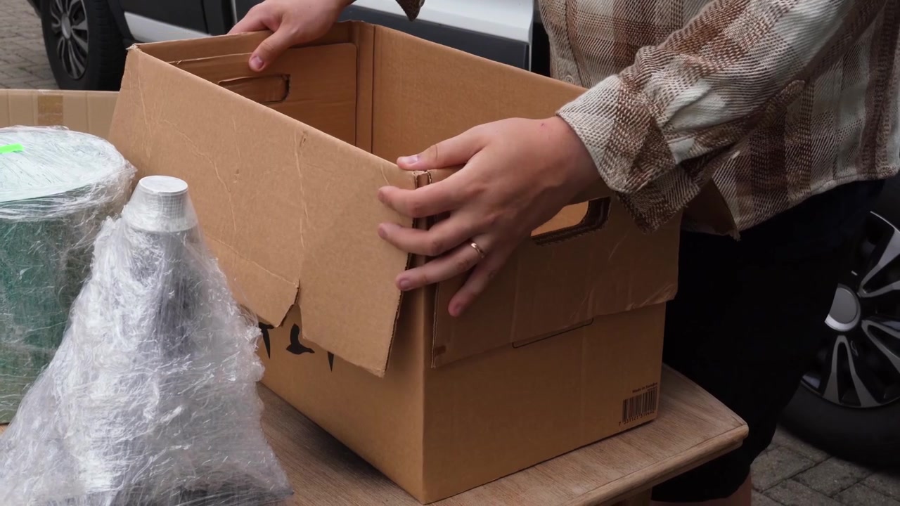 Small business owner packs a cardboard box using reused plastic wrap and old packaging on an outdoor table.