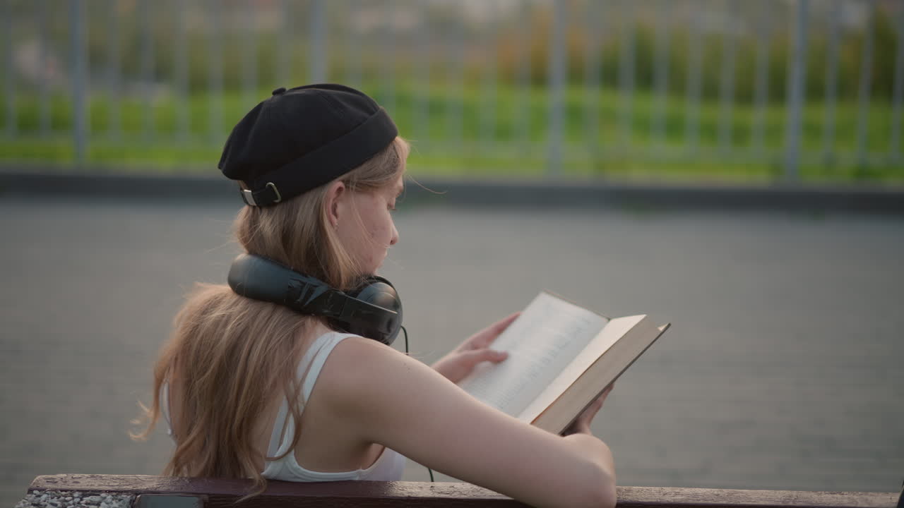 Ung, hvit kvinne som leser bok på en benk i byen i skumringen, iført svart beret og hodetelefoner, blar i en vintage pocketbok, fjernt gjerde og fortau, rolig introspektiv stemning og avslappet livsstil