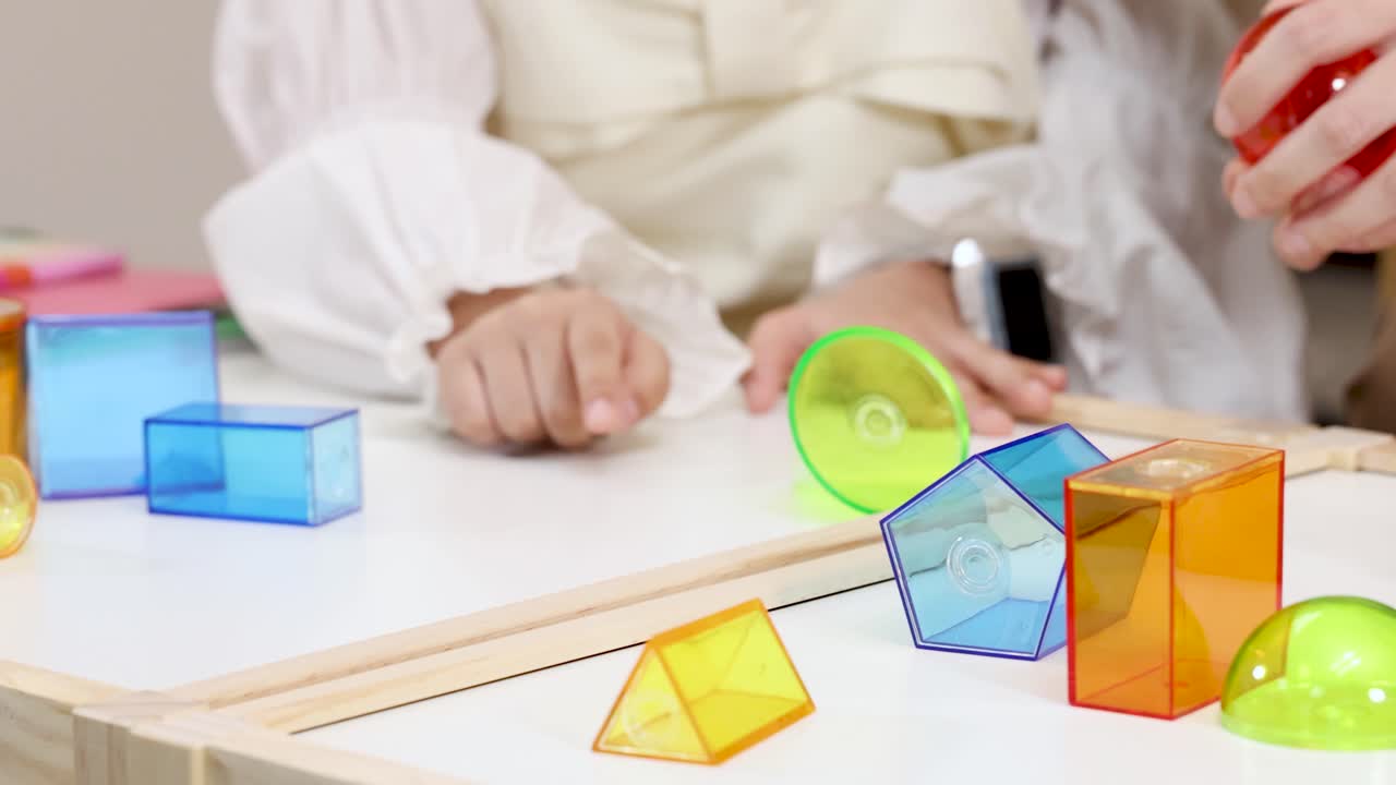 Two people manipulate translucent 3D math shapes on a white table under bright, even lighting