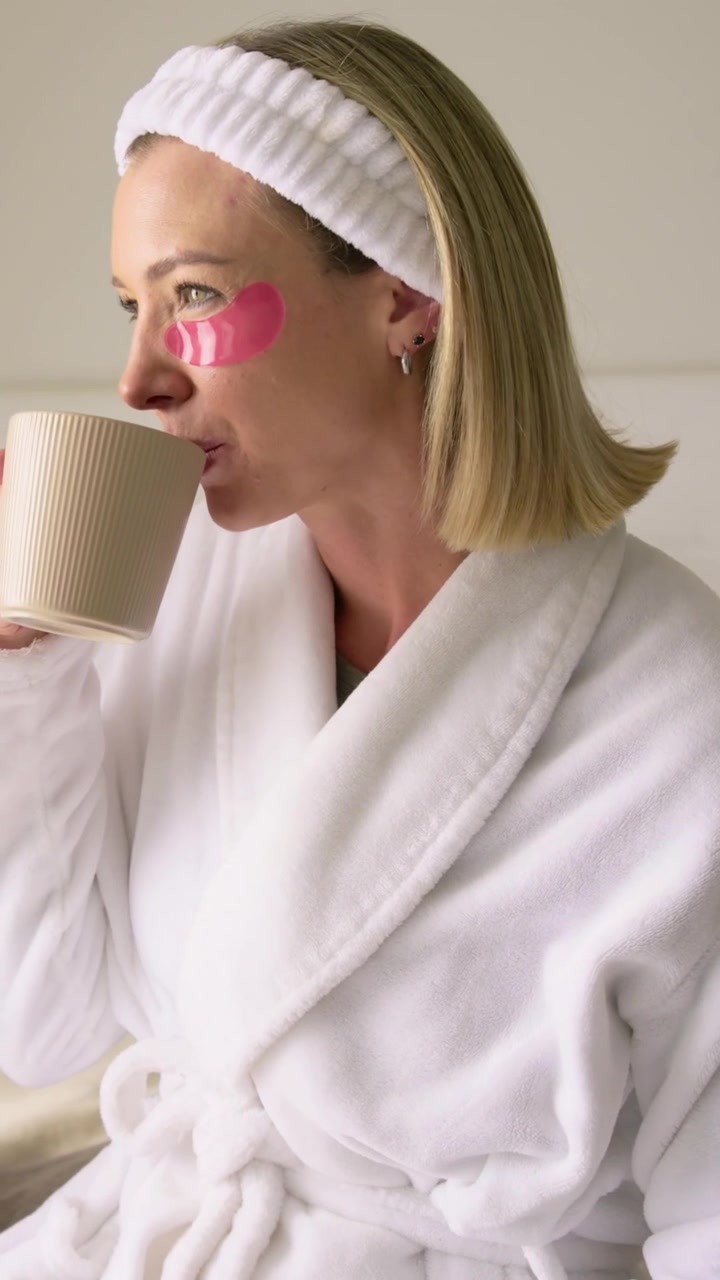 Vertical video: Hearing offscreen call, woman in robe turning, sipping mug on bed, pink patches