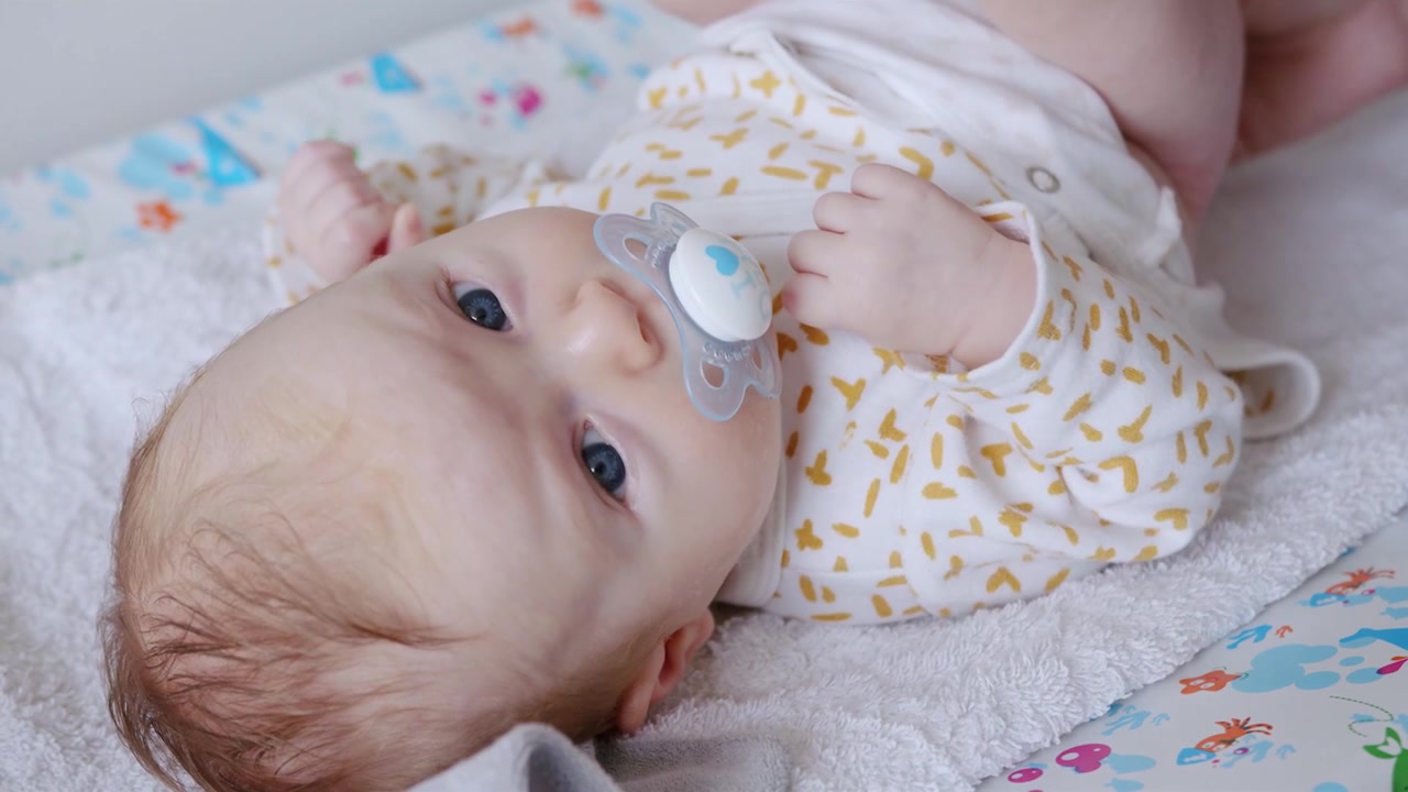 Blonde haired, blue eyed baby stares at the camera sucking a pacifier while her mom cleans her bottom