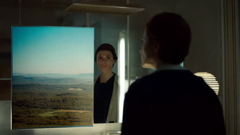woman looking at her reflection in a mirror next to a landscape artwork