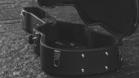 Vista-En-Blanco-Y-Negro-De-Una-Caja-De-Guitarra-Con-Monedas-En-El-Suelo-Y-Un-Hombre-Con-Abrigo-Cantando-Y-Tocando-La-Guitarra-En-La-Calle