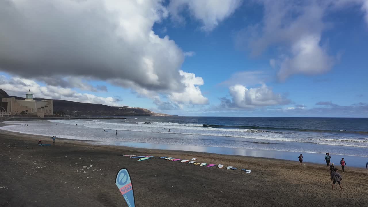 Children’s surf school. Motion lapse left to right. Las Canteras Beach in Las Palmas de Gran Canaria. Blue sky with some clouds. Dark volcanic sand. 4K