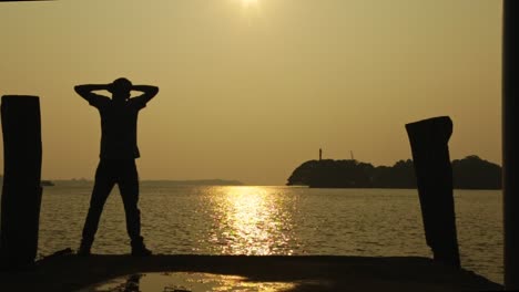 from the edge of the embankment stands a man with outstretched arms , sunset time , houseboats in the background , silhouette