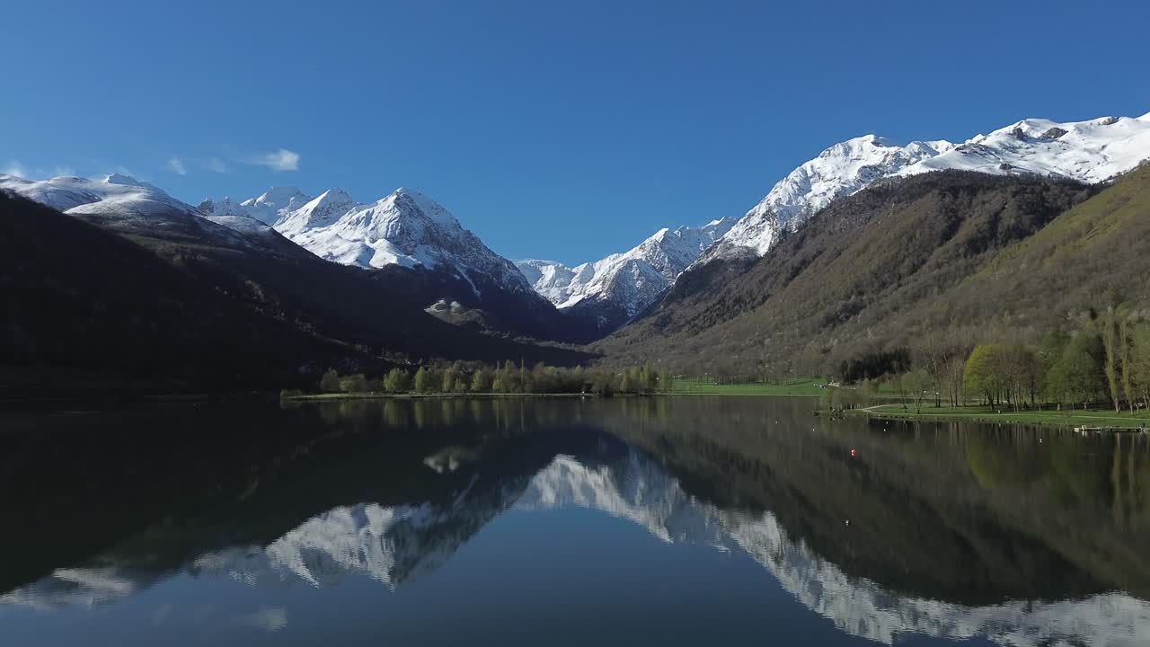 Snow capped mountains mirroring on Lake Génos-Loudenvielle in the French Pyrenees, aerial view with dolly forward and jib up moves.