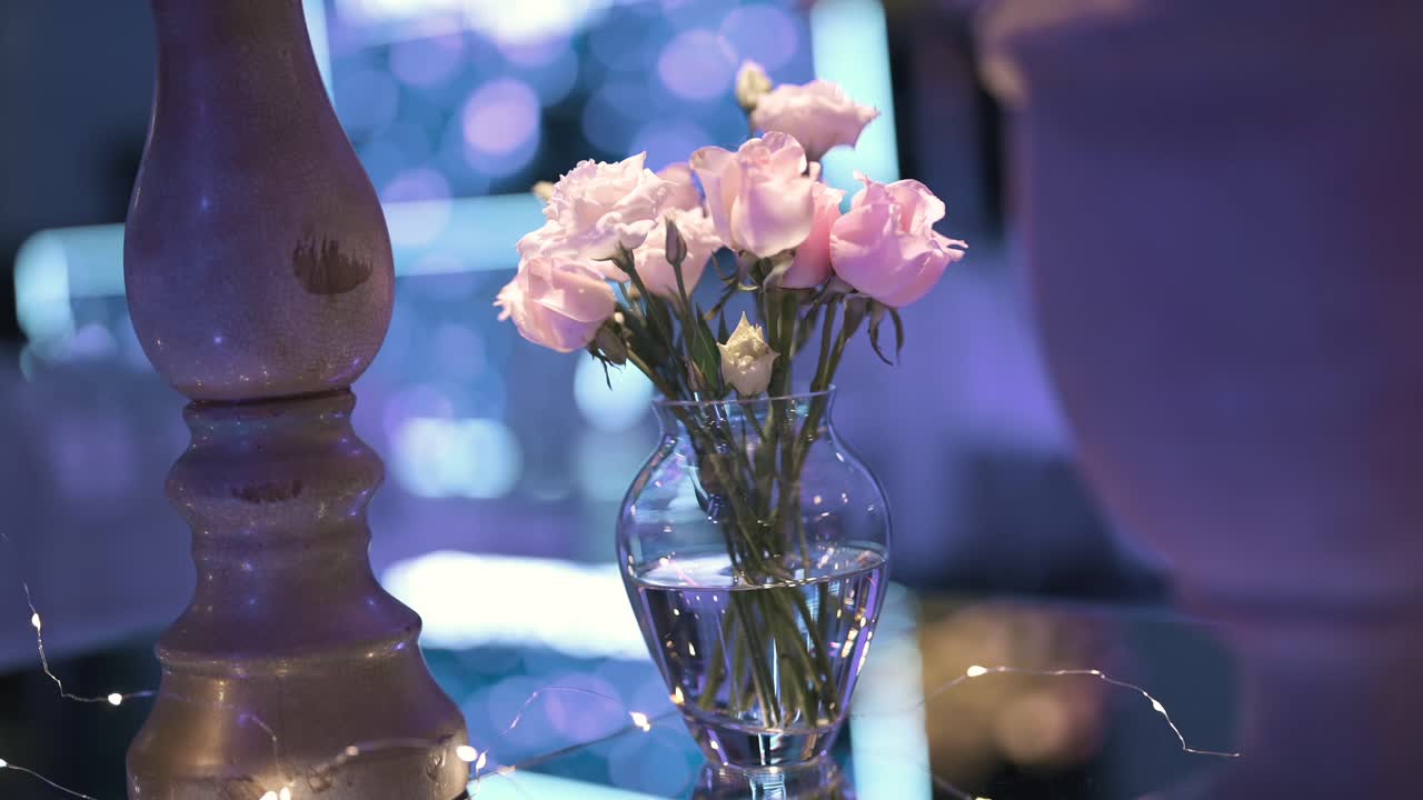 Detailed closeup shot of a luxurious floral arrangement featuring pink and white roses in a transparent glass vase, beautifully illuminated against a blurred wedding reception background