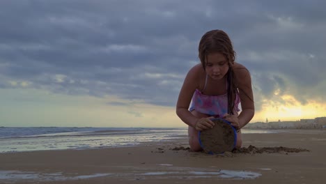 low angle of beautiful caucasian little girl playing with bucket and sand on beach at sunset