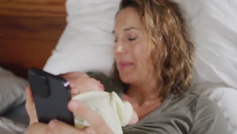 Video-of-happy-caucasian-mother-lying-on-bed-with-sleeping-newborn-baby-and-using-smartphone