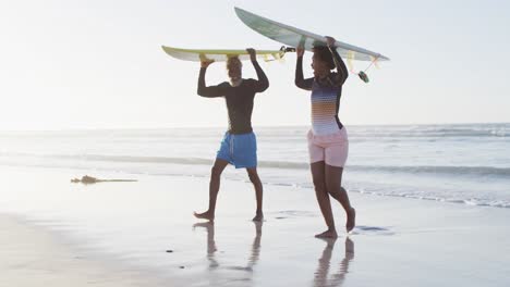 Happy-african-american-couple-walking-with-surfboards-on-sunny-beach