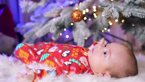 healthy little boy in festive red clothes lies under the christmas tree. baby looking straight ahead. blurred backdrop.