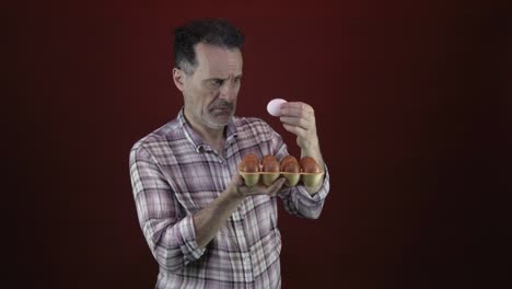 un macho adulto con una mirada desconcertada en su cara examina de cerca un huevo blanco de un cartón de huevos marrones, colocado contra un fondo rojo oscuro