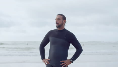 Medium-Shot-Of-A-Bearded-Surfer-In-Wetsuit-Standing-On-Seashore-With-Hands-On-Sides-And-Smiling-At-The-Camera
