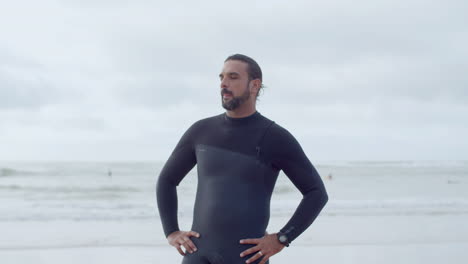 plano medio de un surfista barbudo en traje de neopreno de pie en la orilla del mar con las manos a los lados y sonriendo a la cámara