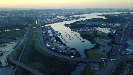 Aufnahmen-Von-Sonnenuntergang-Mit-Blick-Auf-Yachten,-Die-Mit-Verkehr-Auf-Einer-Straße-Festgemacht-Sind,-Und-Schiffscontainern,-Die-Von-Lastwagen-Zu-Schiffen-Transportiert-Werden