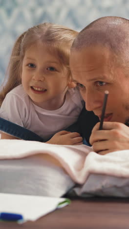 adorable little girl with long ponytails whispers to bald daddy ear lying on floor with soft blanket and pillows in light room staying home