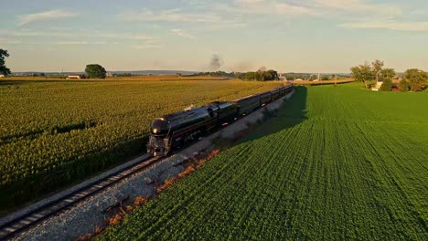 Eine-Luftaufnahme-Einer-Dampfmaschine,-Die-Rauch-Und-Dampf-Bläst,-Mit-Personenwagen,-Die-An-Einem-Schönen-Frühlingstag-Am-Späten-Nachmittag-Durch-Bäume-Und-Ackerland-Auf-Einer-Einzigen-Spur-Durch-Bäume-Und-Ackerland-Fahren