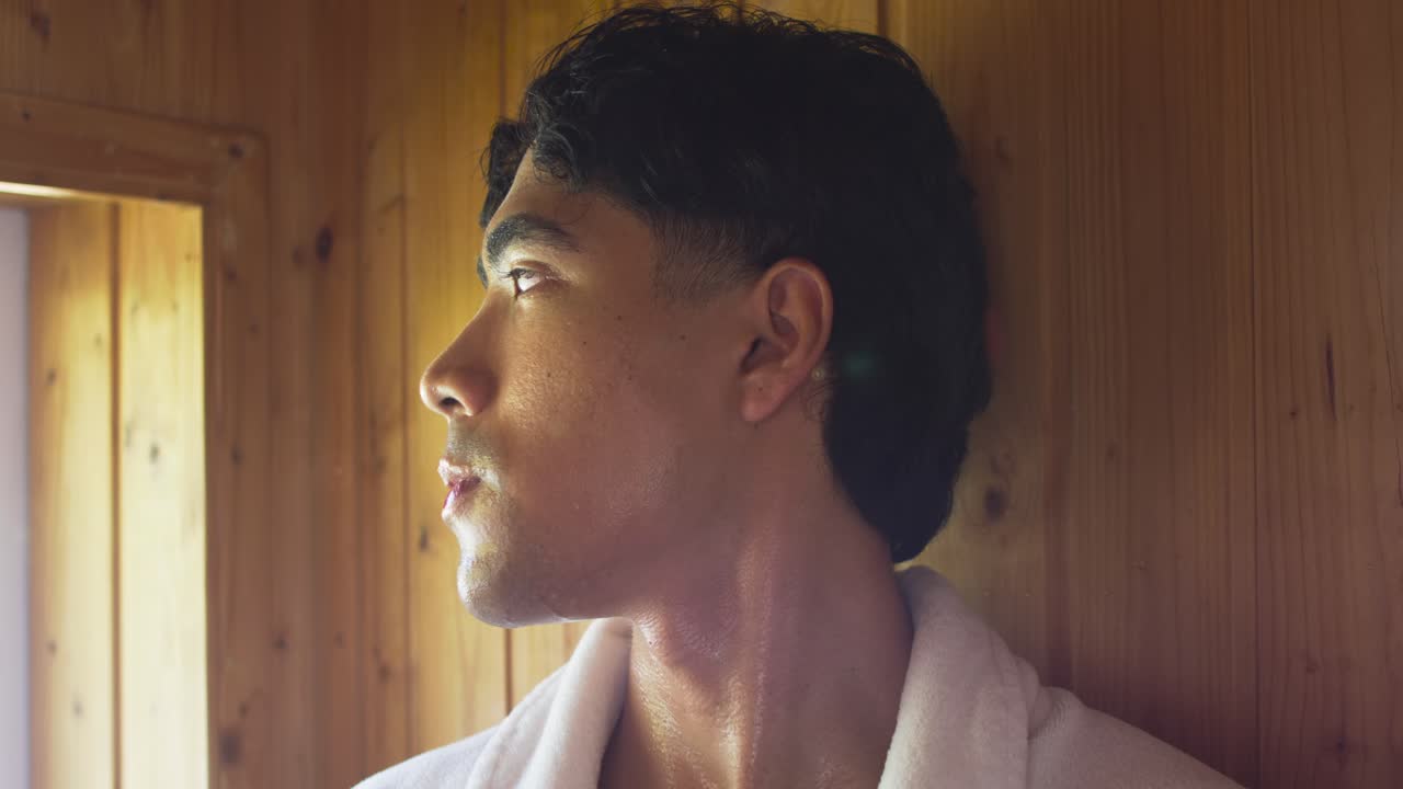 Asiatisk mann i badekåpe sitter mot en trevegg i badstue, reagerer på vinduslys og kikker