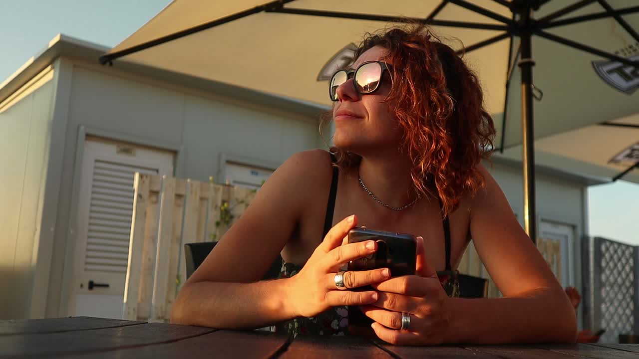 una ragazza dai capelli rossi con gli occhiali da sole si gode il tramonto tenendo in mano uno smartphone