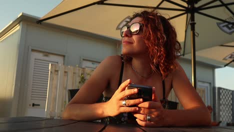 Rothaariges-Mädchen-Mit-Sonnenbrille-Genießt-Den-Sonnenuntergang-Und-Hält-Ein-Smartphone-In-Der-Hand