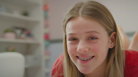 Teenage-girl-wearing-orthodontic-braces-lying-on-bed-at-home-turns-to-face-camera-and-smiles---shot-in-slow-motion