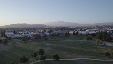 A-drone-glides-gently-over-a-park-amidst-a-sea-of-commercial-warehouses,-bathed-in-the-warm-golden-hues-of-a-setting-sun,-capturing-a-serene-and-captivating-evening-landscape