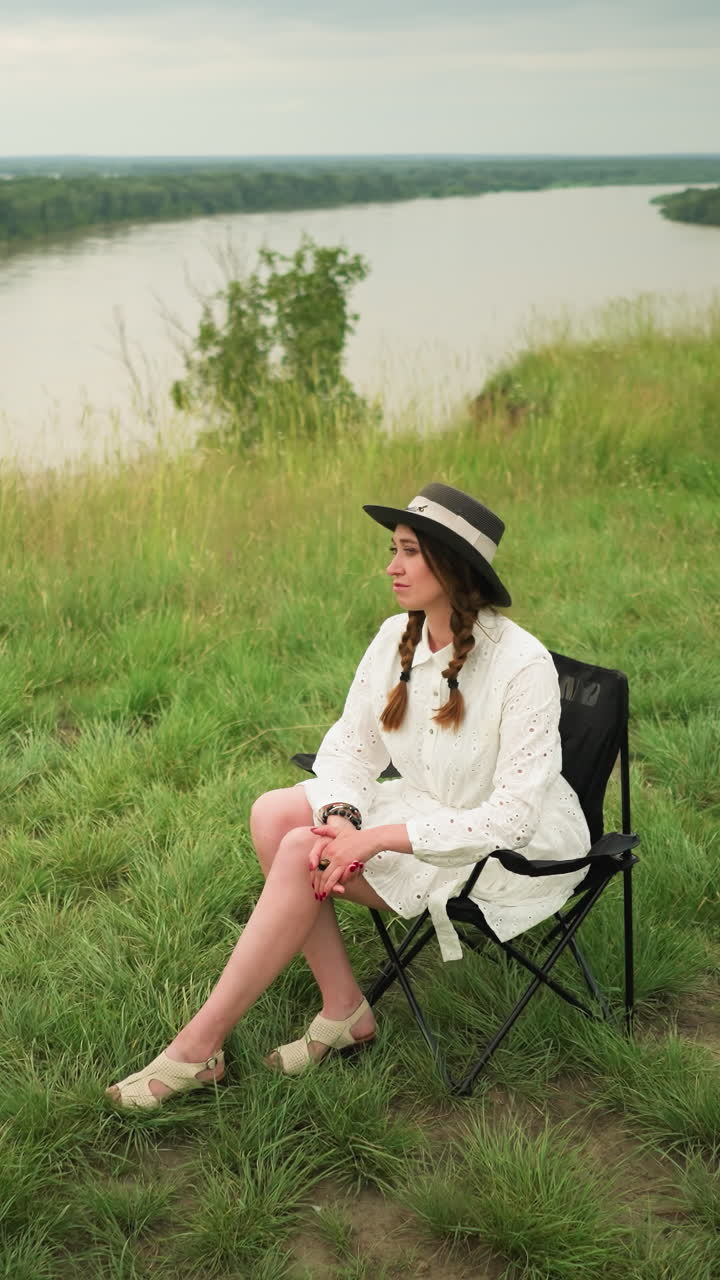 세련된 모자와  드레스를 입은 우아한 여성이 조용한 호수 의 울창한 풀에서 의자에 앉아 있습니다. 그녀는 우아하게 포즈를 취하고 구름이 가득한 하늘 아래 평화로운 순간을 포착합니다.