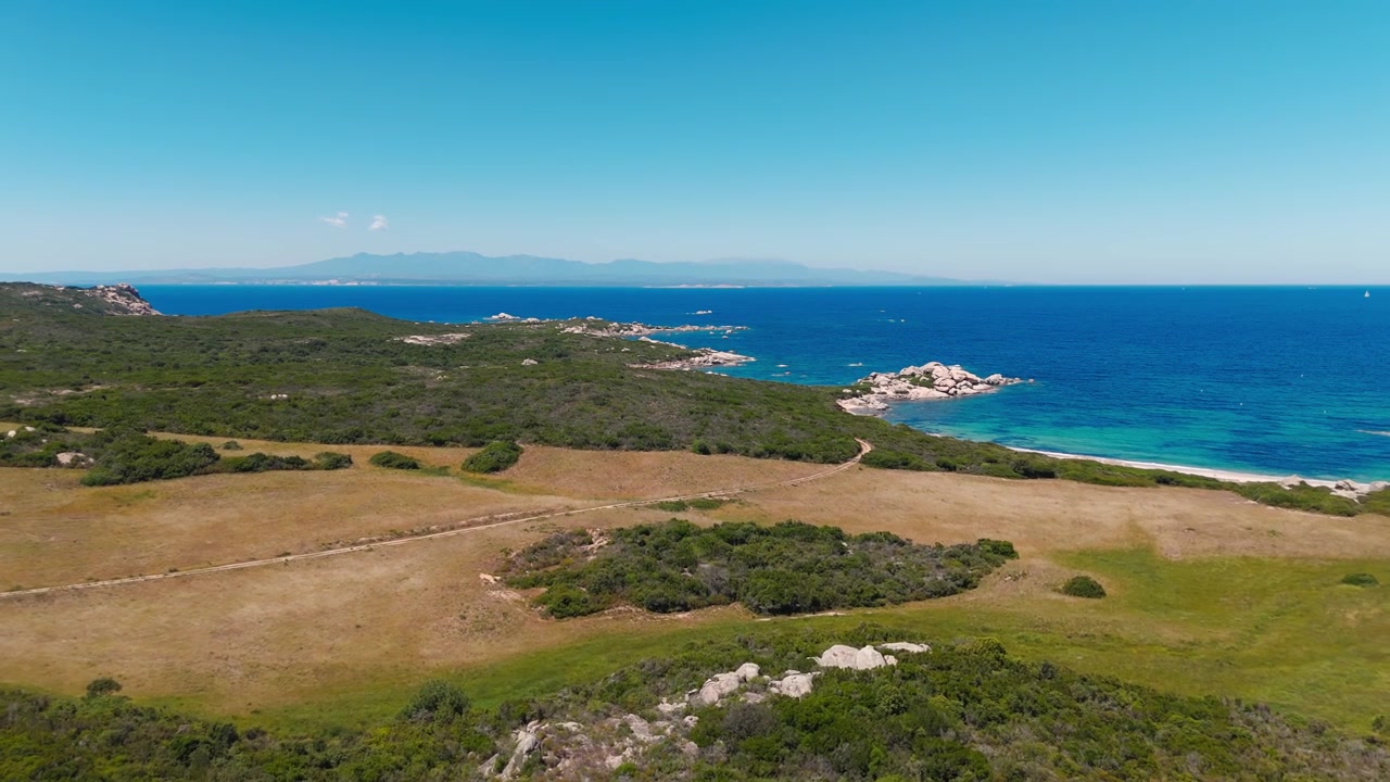 Vista aerea della costa sarda con mare cristallino, costa rocciosa e campi aperti.