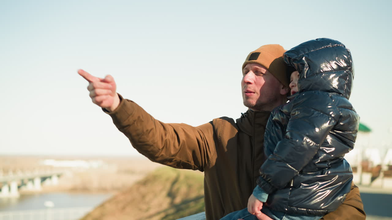 un padre lleva a su hijo mientras está de pie cerca de una vista panorámica, el padre, con una chaqueta y sombrero marrones, está explicando algo al niño, que está vestido con una jaqueta negra
