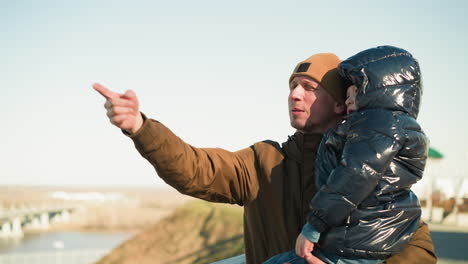 a father carries his son while standing near a scenic overlook, the father, wearing a brown jacket and hat, is explaining something to the child, who is dressed in a black jacket