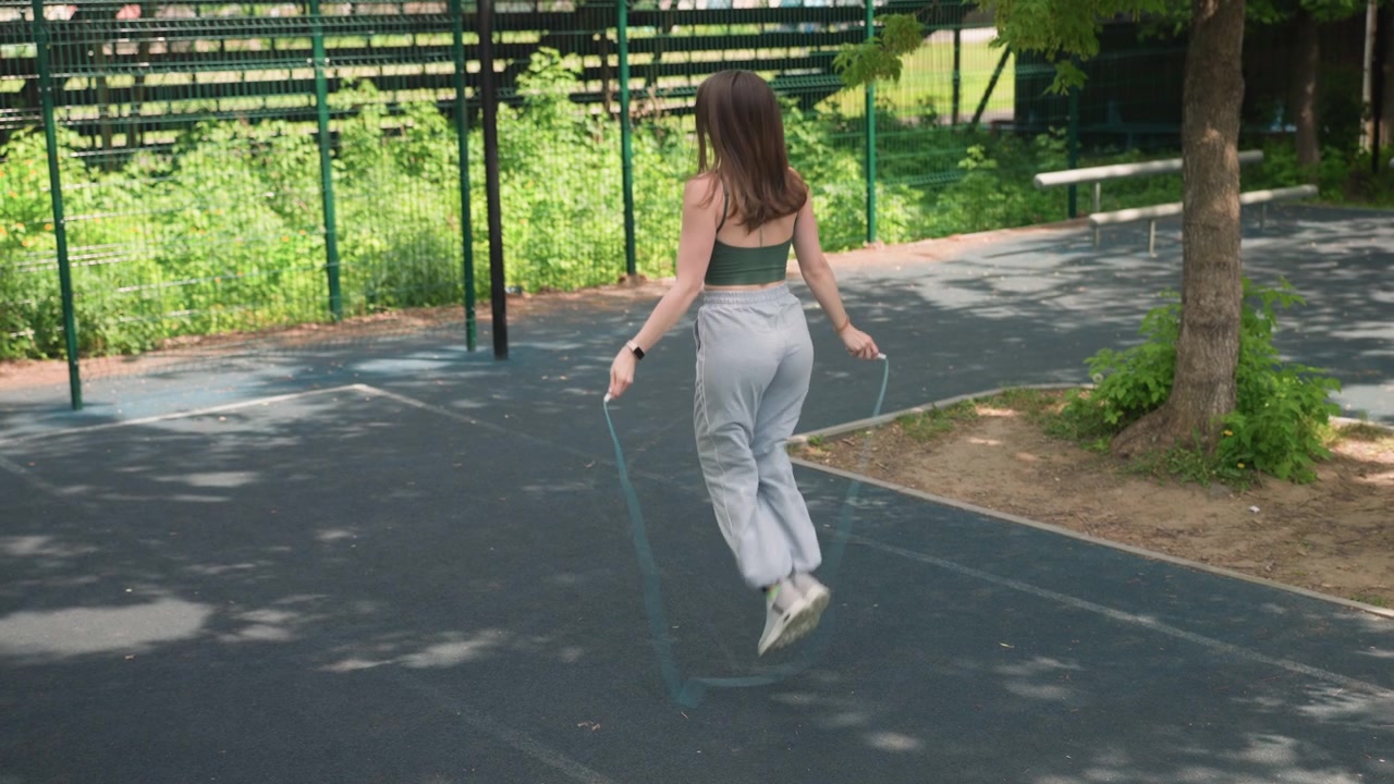 Close up of young woman exercising by skipping rope on outdoor court. Woman wears casual sportswear and maintains steady motion with consistent natural lighting and greenery in background