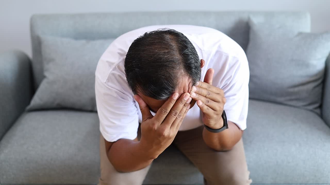 Uomo asiatico stanco e frustrato che si tocca la testa, con mal di testa, emicrania, seduto sul divano di casa, nonno solo e triste che affronta la perdita familiare, il dolore, un brutto trauma, un esaurimento nervoso, una crisi di età