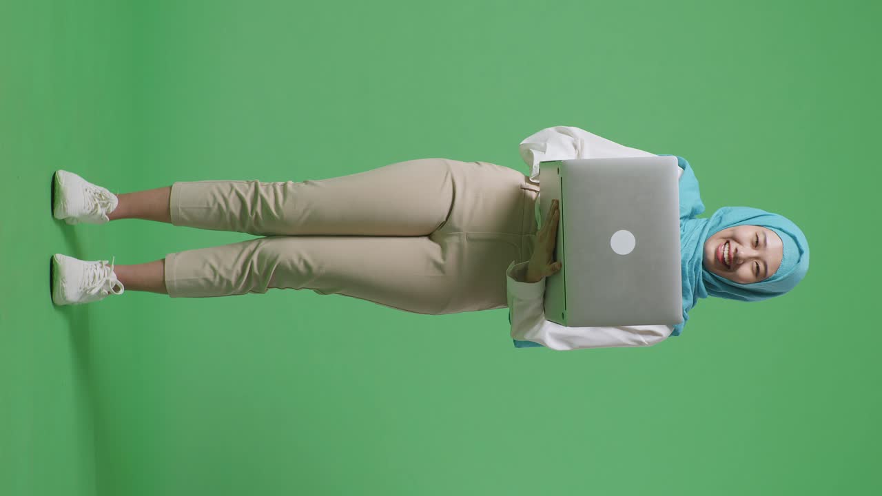 cuerpo lleno de mujer musulmana asiática escribiendo en una computadora portátil y sonriendo a la cámara mientras está de pie en el estudio de fondo de la pantalla verde