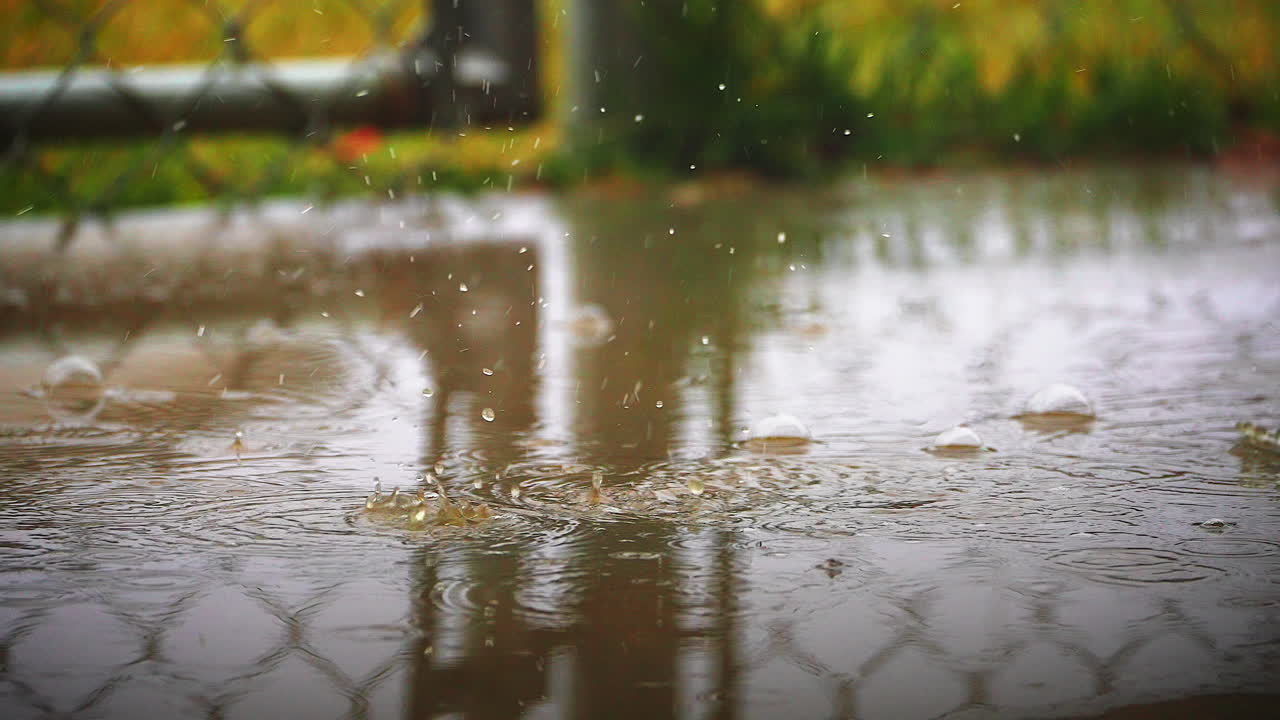 Raindrops falling from the sky and splashing in a backyard puddle by a chain-link fence or gate - slow motion