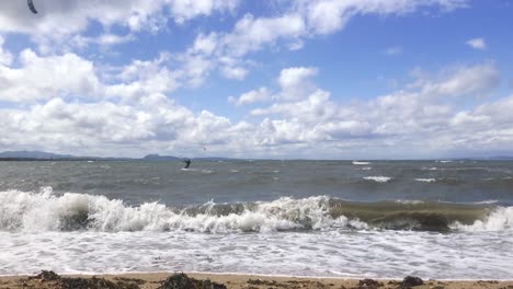 A-fixed-shot-of-waves-breaking-on-a-sandy-beach-with-kite-surfers-moving-through-the-shot-in-the-background,-on-a-windy-summer-day-|-Portobello-beach,-Edinburgh-|-Shot-in-HD-at-cinematic-24-fps