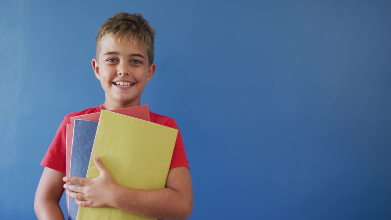 Porträt eines glücklichen kaukasischen Jungen mit Büchern auf blauem Hintergrund, Kopierraum, Zeitlupe.