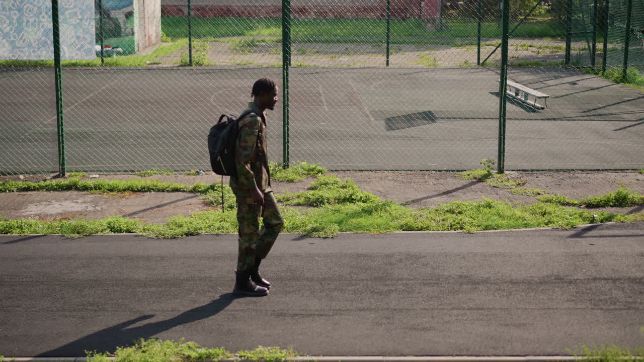 Soldier Walks Away, Service Member Strolling Along Fence With Gear Under Sunlight, Soldier In Camouflage Suitcase Walks Past Fence With Elongated Shadow Indicating Solitude And Determination