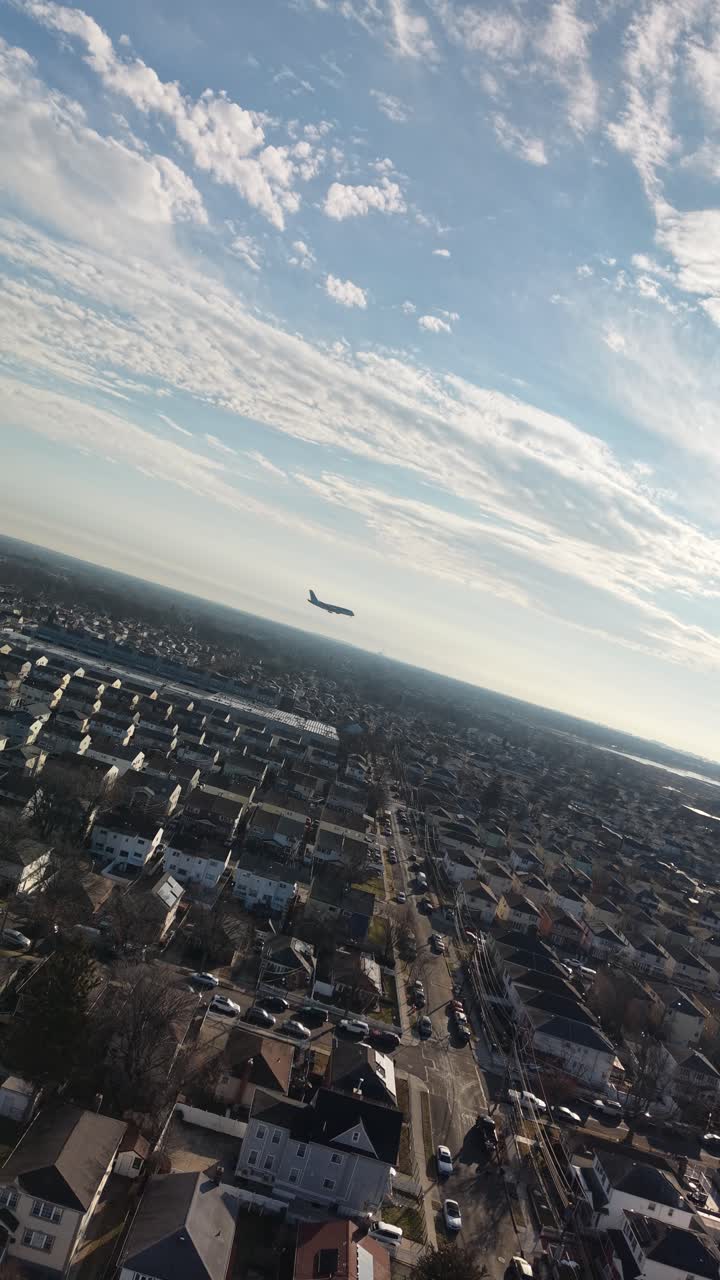 Dronebillede i Queens, New York af et passagerfly, der flyver over et beboelsesområde i byen under en smuk solopgang. Klar sol, der skinner gennem spredte skyer på himlen