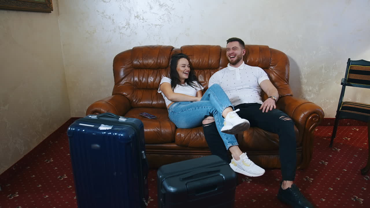 Joyful young couple on a couch. Couple in love having happy time together before leaving the hotel. Travel suitcases in the hotel room.