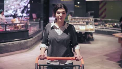 Young-beautiful-brunette-girl-smiles-enjoying-walking-in-grocery-store-with-push-cart.-Shopping-in-a-supermarket