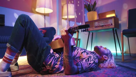 young stylish caucasian man with long hair lying on the floor and playing the guitar in a retro party at home