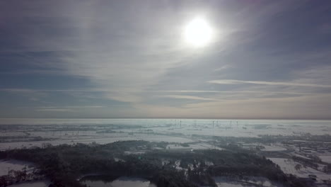 Aerialshot-of-snowy-winter-landscape-with-forest-trees-and-wind-turbine-farm-in-background-during-sunlight