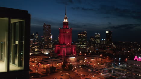 Panoramic-view-of-square-and-red-lit-Palace-of-Culture-and-Science-at-night.-Down-town-skyscrapers-in-background.-Warsaw,-Poland