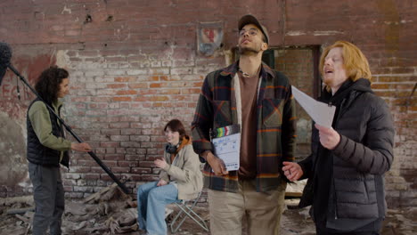 dos compañeros de producción hablando y leyendo un documento sobre la película en un edificio en ruinas mientras otros compañeros hablan y esperan para comenzar la grabación