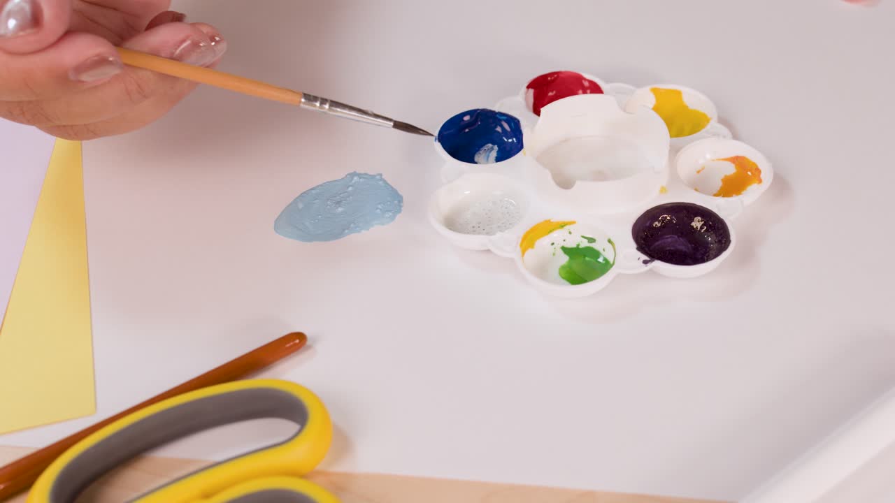 Artist hand mixes blue and white watercolor paints on palette under bright, even overhead lighting