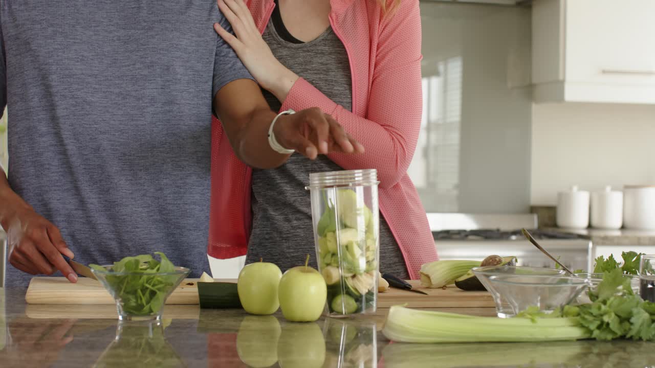 Un hombre está cortando pepino sobre la encimera de la cocina después de que una mujer señale una batidora para hacer un batido verde