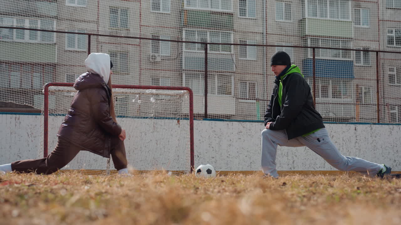 골대 근처에서 스트레칭하는 백인 여성 선수와 슛을 준비하는 백인 남성 선수, 추운 도시의 경기장, 편안한 겨울옷, 장난기 넘치는 긴장감, 가까운 자세와 시선 교환, 동네 스포츠
